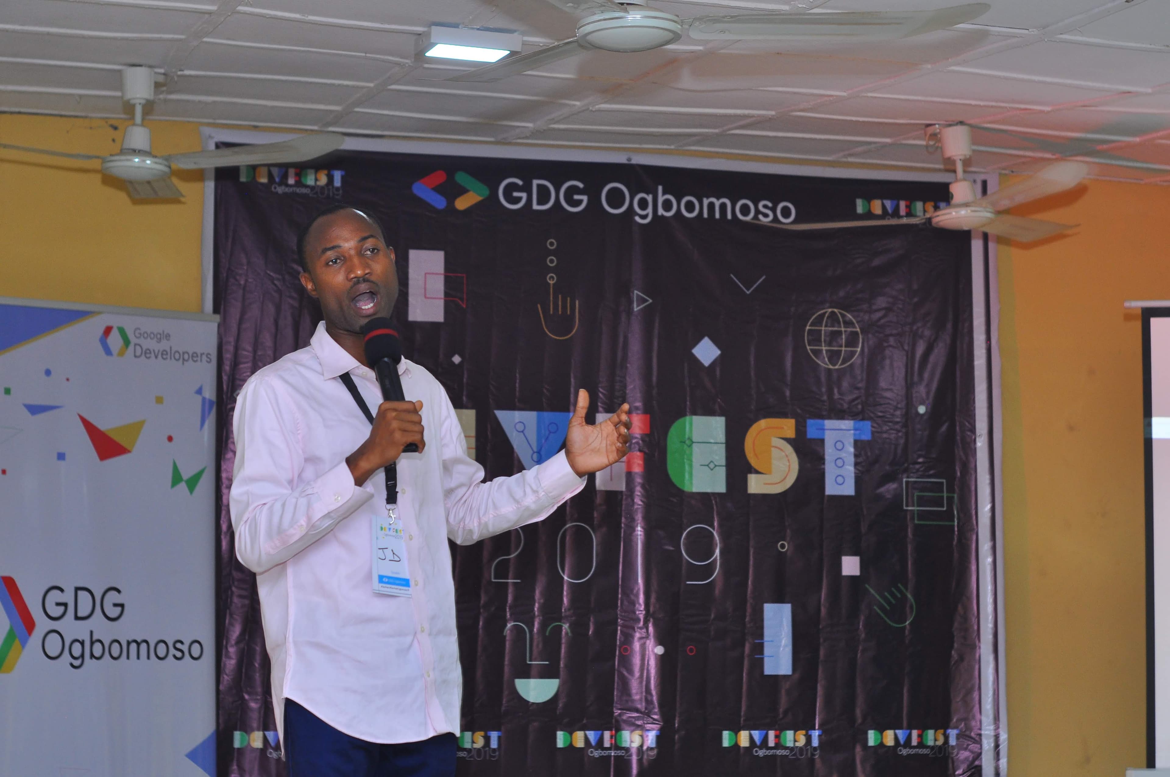 A speaker in a pink shirt with a microphone by the GDG backdrop at DevFest Ogbomoso 2019