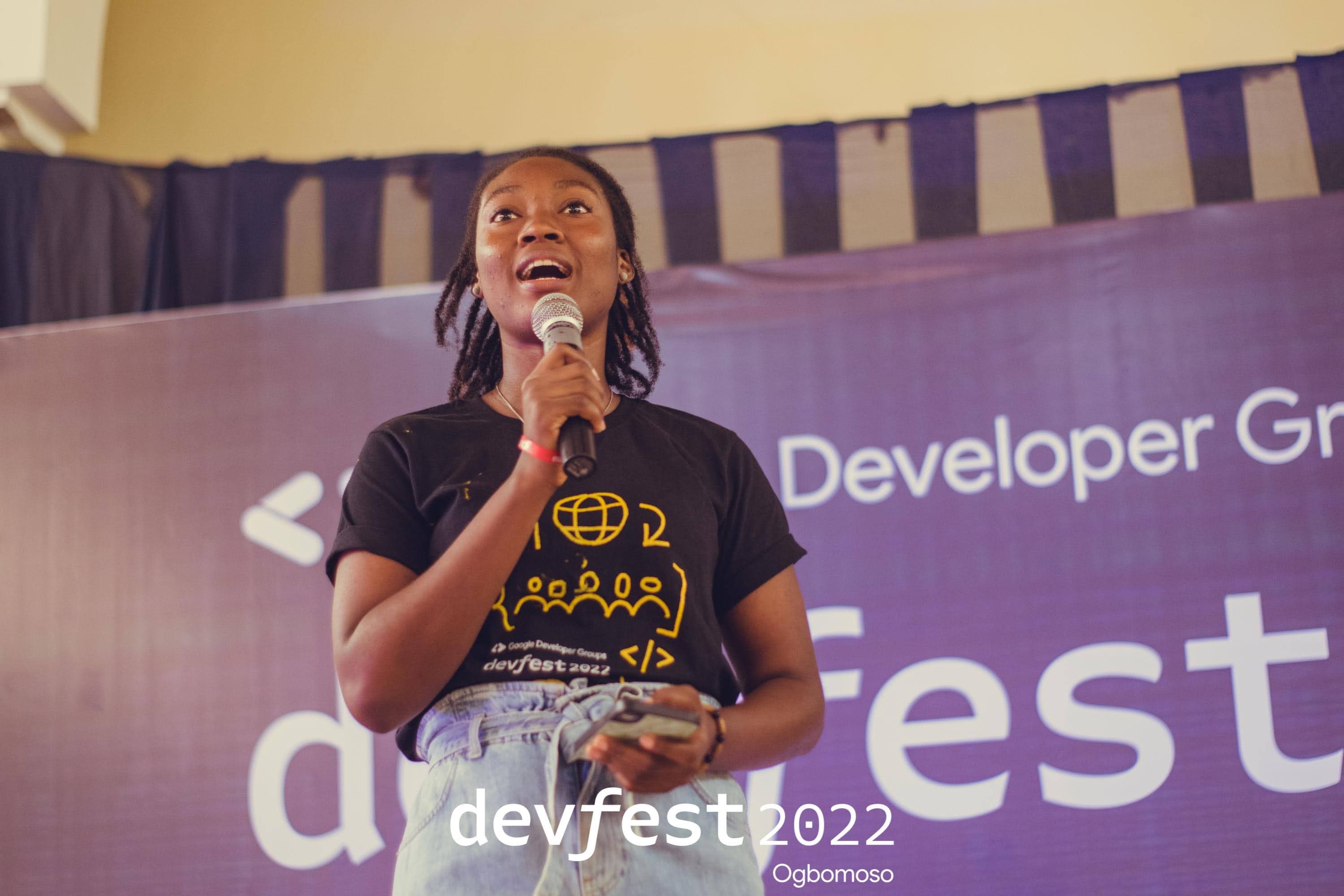 A person speaking into a microphone in front of the DevFest banner at DevFest Ogbomoso 2022