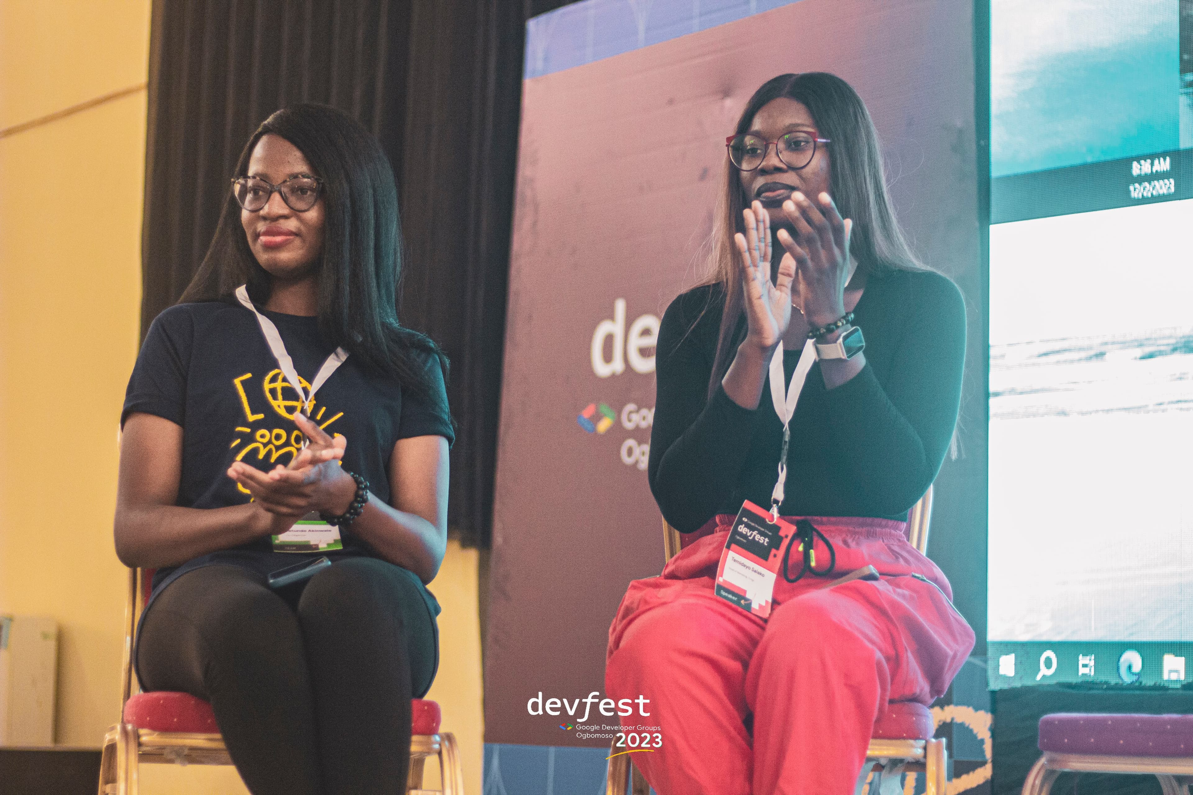 Two people seated on stage, clapping beside a screen at DevFest Ogbomoso 2023