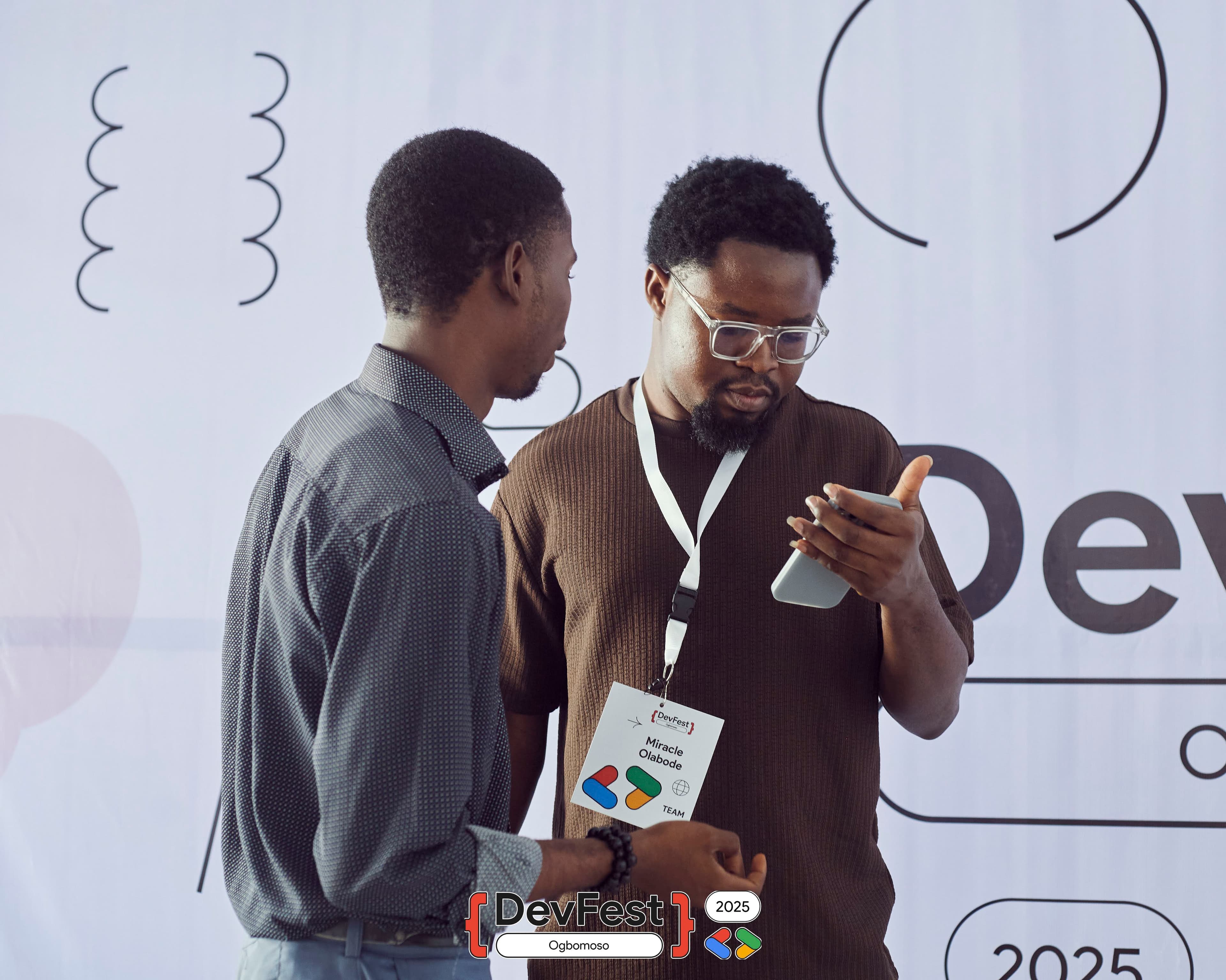 Two people by the DevFest backdrop, one checking a phone at DevFest Ogbomoso 2025