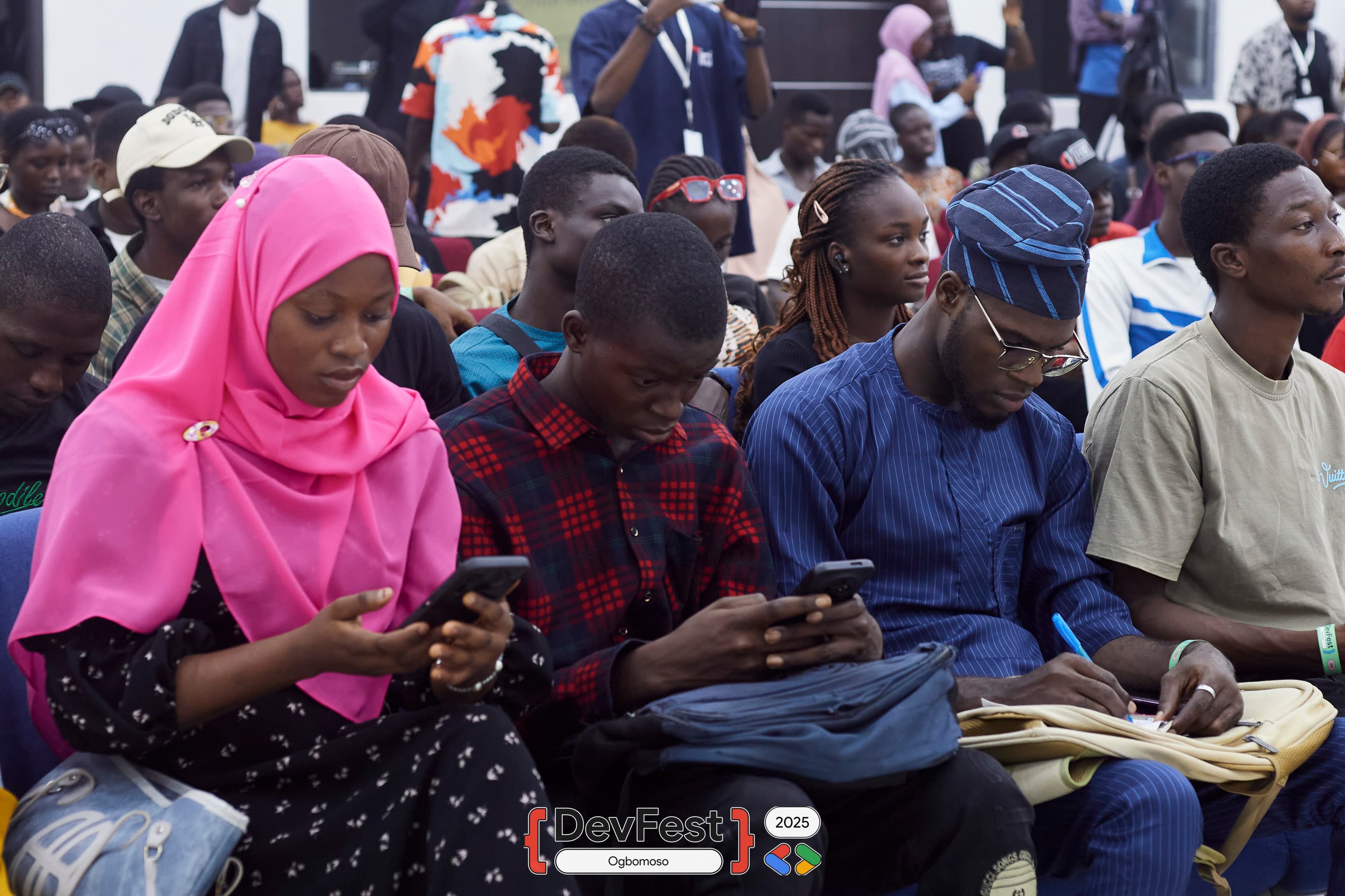 Seated attendees on phones while one writes notes at DevFest Ogbomoso 2025